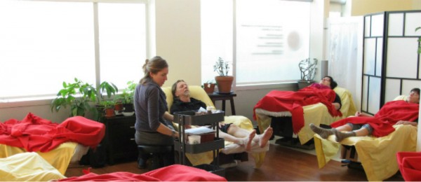 Community acupuncture practitioner sitting on a stool in the GCA group treatment room, in front of a bearded patient relaxing in a recliner, while several other patients sleep in recliners covered in red blankets in a loose circle in the room.