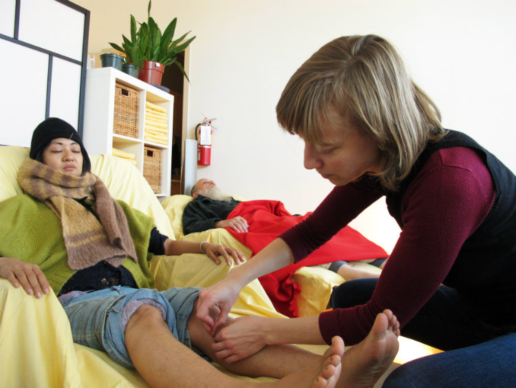 A women sets needles into a patient's leg while patient reclines with closed eyes in a recliner. Another patient sleeps in another in the background.