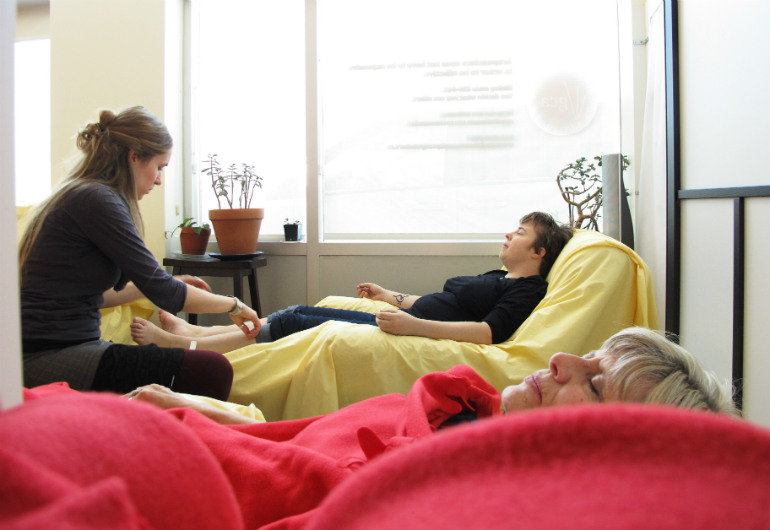 A woman sleeping under a red blanket in the foreground; in the background a practitioner treating a patient in a black shirt reclining on a lazyboy by two jade plants.