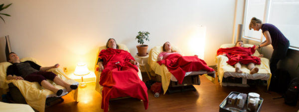 Four people reclined in lazyboys under red blankets with practitioner standing to the right, adjusting a blanket