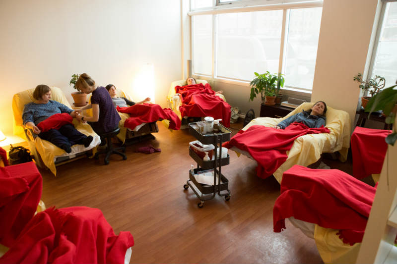 9 recliners in a semi-circle, each with a patient resting under red blankets. Practioner seated on a small black stool giving a treatment to one patient.