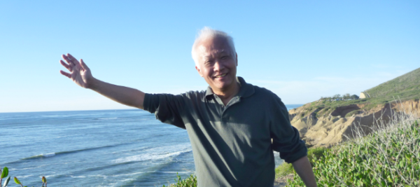 A balding man stands in the sunshine with a huge smile waves to the camera. Ocean and rugged coastline behind him.