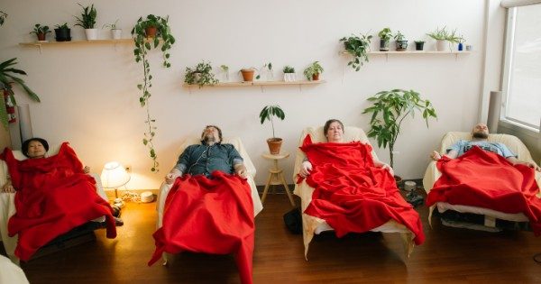 Four acupuncture patients sleeping under red blankets in four recliners lined up against a wall lined with plants.