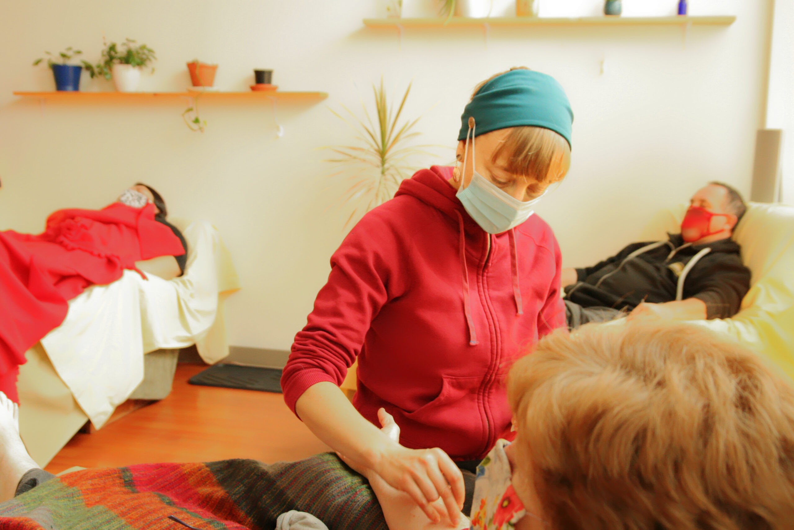 A person in a surgical mask and a red hoodie sets needles into someone's arm. Visible in the background are two other masked patients with closed eyes and two shelves on the wall holding plants.