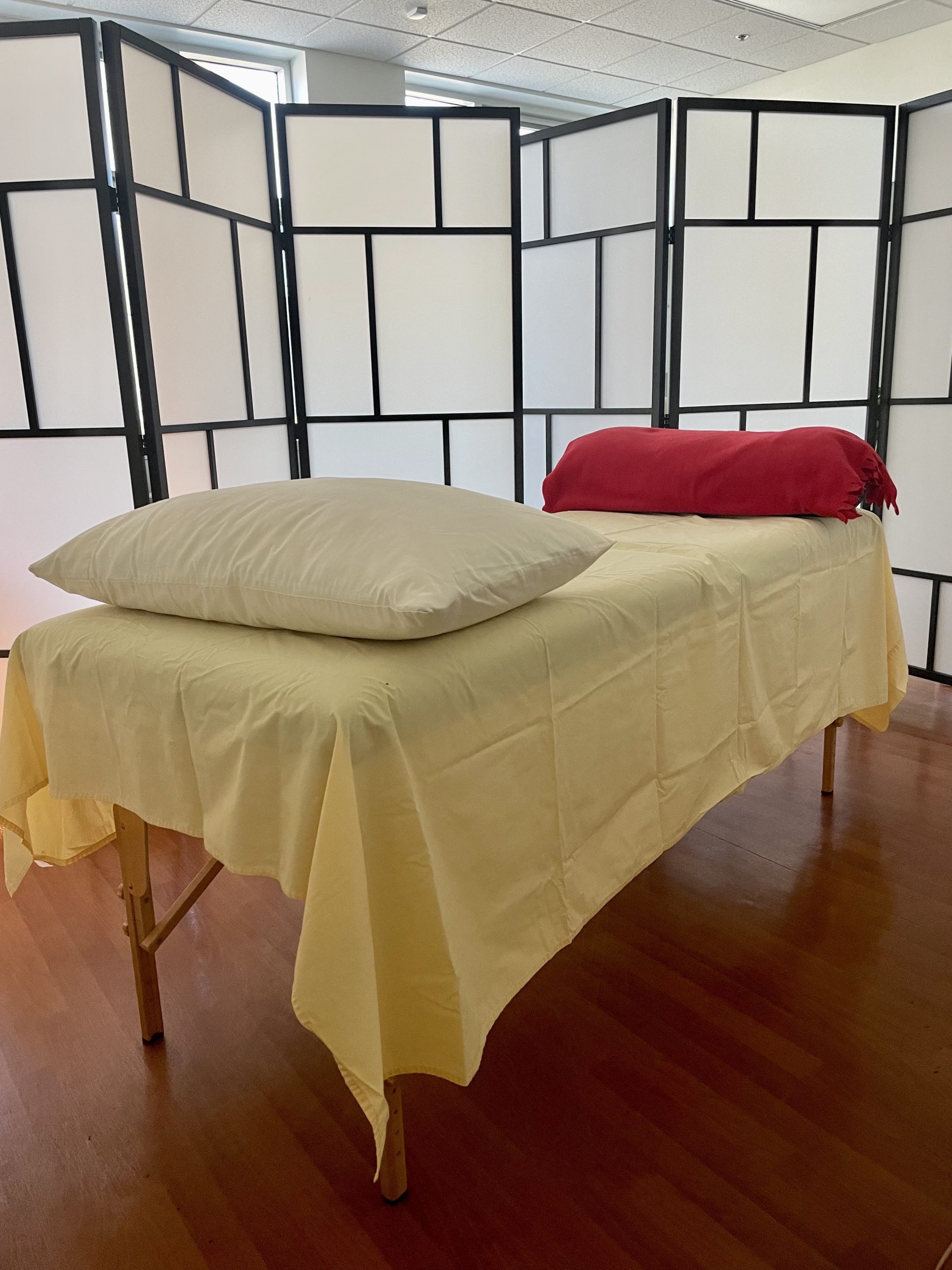 Massage table draped in a yellow sheet with a white pillow on the near end and a red bolster on the far end. View is on an angle. White and black screens are visible in the background.