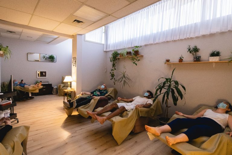 Four masked people lying back in recliners in a softly lit room with many plants.